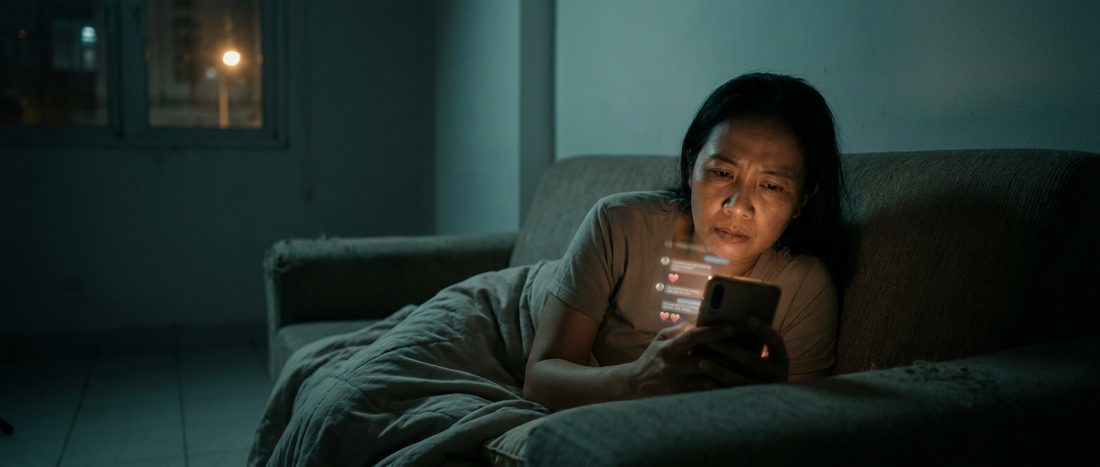 A woman on a sofa at night reading a long chat thread by phone light